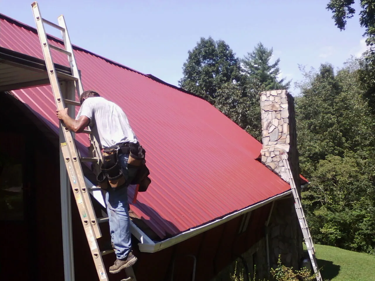 Roofer on a ladder beside a red metal roof and stone chimney — Soffit & Fascia Repair by Roofing Brothers in Salida
